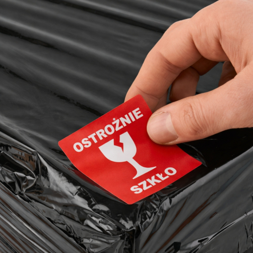 dłoń naklejająca naklejkę kurierską "nie rzucać" na karton owinięty czarną folią stretch